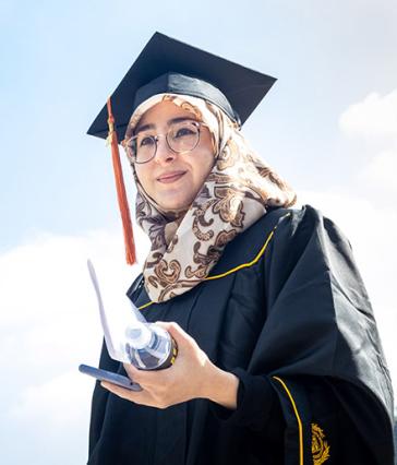Student wearing hijab and graduation gap holds notepad and water bottle